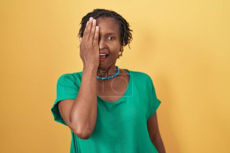 Téléchargez les photos : Femme africaine avec dreadlocks debout sur fond jaune couvrant un oeil avec la main, sourire confiant sur le visage et l'émotion surprise. - en image libre de droit