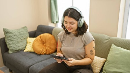 Téléchargez les photos : Jeune femme hispanique souriante jouissant de musique sur un casque, assise sur un canapé à la maison. - en image libre de droit