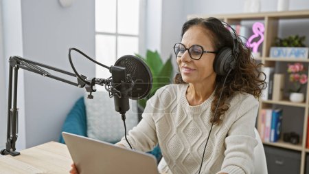 Téléchargez les photos : Une femme hispanique mature souriante portant un casque parle dans un microphone de studio tout en lisant depuis une tablette à l'intérieur. - en image libre de droit
