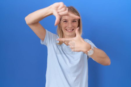 Foto de Mujer caucásica joven con camiseta azul casual sonriendo haciendo marco con las manos y los dedos con la cara feliz. concepto de creatividad y fotografía. - Imagen libre de derechos