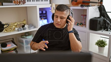 Foto de Apasionado joven músico latino inmerso en una enérgica actuación musical, usando hábilmente la computadora y los auriculares en su estudio de música, canalizando su arte en el interior - Imagen libre de derechos