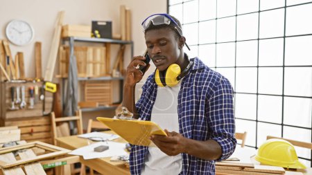 Foto de Hombre afroamericano al teléfono en un taller de carpintería mirando portapapeles - Imagen libre de derechos
