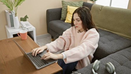Téléchargez les photos : Une jeune femme travaille sur son ordinateur portable dans un salon confortable, présentant un style de vie moderne à la maison. - en image libre de droit
