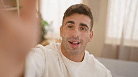 Photo for Cheerful young hispanic man sitting on bed, engaging in a heartwarming digital conversation via video call. captured in a cozy bedroom, showing the blend of modern technology, and home comfort. - Royalty Free Image