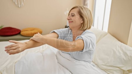 Foto de Una mujer rubia de mediana edad se aplica loción en el brazo en un dormitorio bien iluminado, capturando una rutina personal de cuidado de la piel. - Imagen libre de derechos