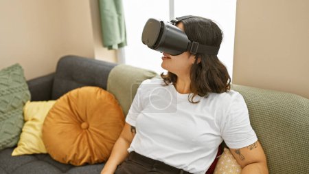 Foto de Mujer hispana joven disfruta de auriculares de realidad virtual en el acogedor entorno de la sala de estar - Imagen libre de derechos
