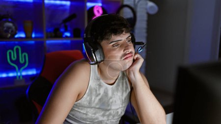Téléchargez les photos : Un jeune homme ennuyé avec des écouteurs dans une salle de jeux éclairée au néon regardant un écran d'ordinateur la nuit. - en image libre de droit