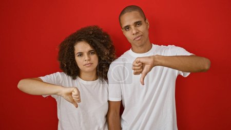 Hermosa pareja expresando negación con un gesto de pulgar hacia abajo sobre un fondo rojo aislado