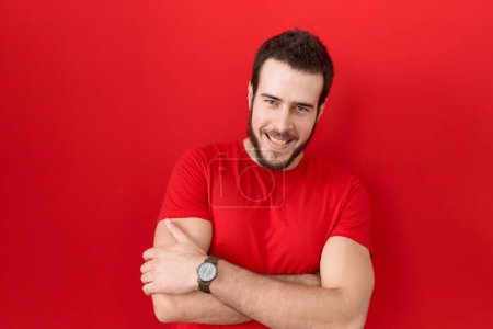 Téléchargez les photos : Jeune homme hispanique portant un t-shirt rouge décontracté heureux visage souriant avec les bras croisés regardant la caméra. personne positive. - en image libre de droit