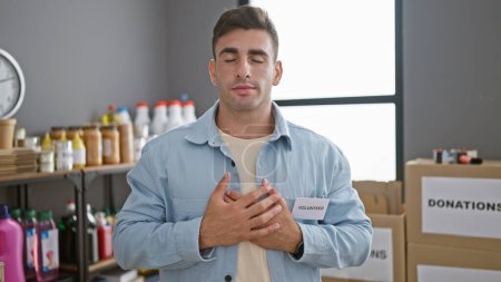 Emotional portrait of a handsome young hispanic man, volunteering at a charity center, standing with hands on chest, making a heart gesture in a calm and serious air, indoors at the warehouse.