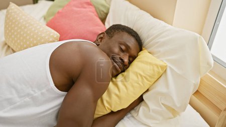Foto de Hombre afroamericano que duerme tranquilamente en un dormitorio luminoso y colorido con almohadas y luz solar. - Imagen libre de derechos