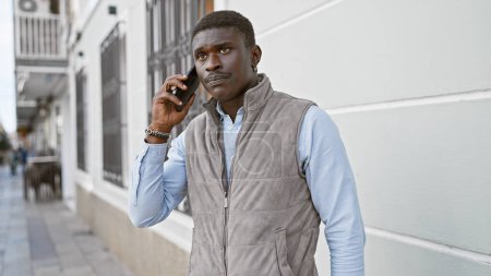 Un homme africain en gilet utilise un téléphone portable dans une rue de la ville, respirant la confiance et le style.