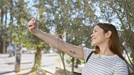 Foto de Una joven sonriente con un tatuaje se toma una selfie en un parque soleado con exuberante vegetación. - Imagen libre de derechos