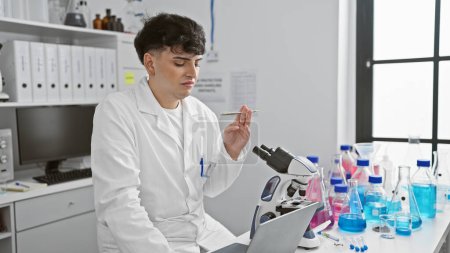 Photo for A young man in a white lab coat examines a pen while working with a microscope in a well-equipped laboratory. - Royalty Free Image
