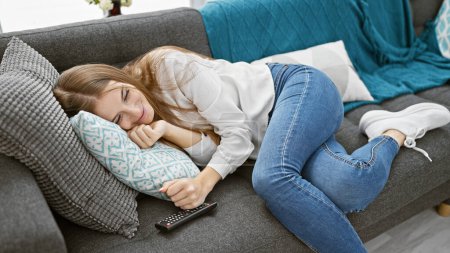 Téléchargez les photos : Jeune femme détendue se prélassant sur un canapé dans un salon confortable, souriant à l'aise avec télécommande au premier plan. - en image libre de droit