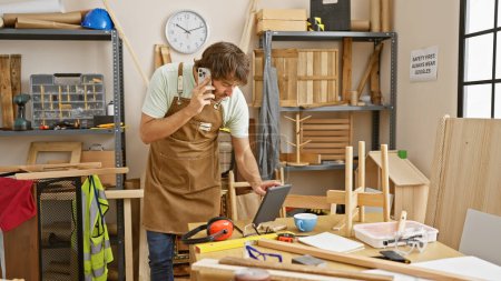 Téléchargez les photos : Un homme barbu dans un atelier discute sur son téléphone tout en naviguant sur une tablette au milieu des outils de travail du bois. - en image libre de droit