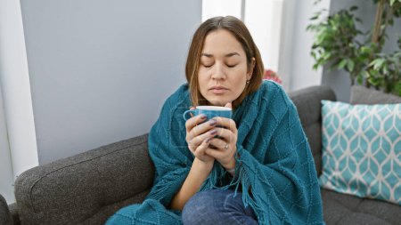 Photo for A relaxed woman enjoys a warm drink on a sofa in a cozy living room, wrapped in a teal blanket. - Royalty Free Image