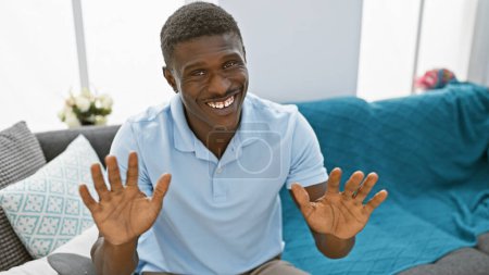 Téléchargez les photos : Homme afro-américain souriant vêtu avec désinvolture dans un salon avec canapé et oreillers décoratifs. - en image libre de droit