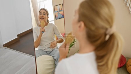 Foto de Una mujer rubia de mediana edad canta juguetonamente en un cepillo como un micrófono en un acogedor entorno de sala de estar. - Imagen libre de derechos