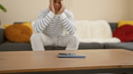 Photo for Worried young hispanic man waits nervously for a smartphone call, sitting home on the sofa, a portrait of anxiety in his handsome expression. - Royalty Free Image