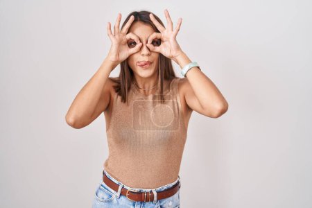 Téléchargez les photos : Young hispanic woman standing over white background doing ok gesture like binoculars sticking tongue out, eyes looking through fingers. crazy expression. - en image libre de droit