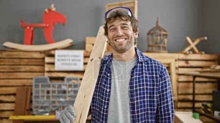 Handsome young man with beard, skilled carpenter, a pro in his business, smiling behind glasses. holds lumber plank in carpentry workshop, ready to conquer woodworking industry!