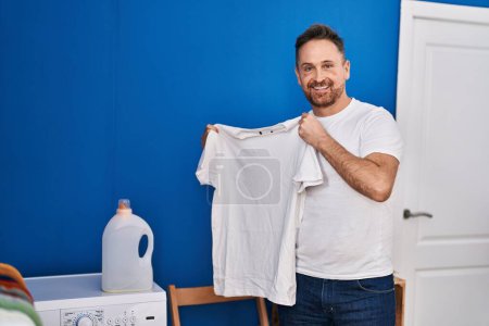 Photo for Young caucasian man smiling confident holding clean t shirt at laundry room - Royalty Free Image