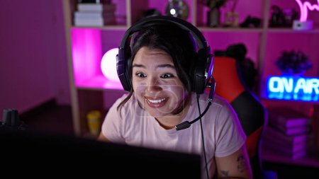 Smiling young hispanic woman with headphones in a vibrant gaming room at home