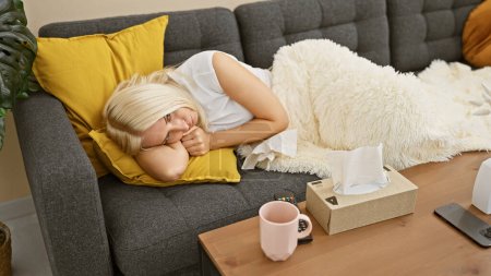 Mujer joven enferma, una hermosa rubia, acostada en el sofá en casa, a distancia en la mano, absorta en la televisión, luchando contra los síntomas de la gripe mientras se acurrucaba bajo una manta.