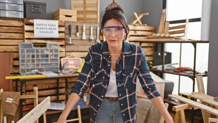 Téléchargez les photos : Une femme mûre portant des lunettes de sécurité se tient en confiance dans un atelier de menuiserie, entourée d'outils et de bois. - en image libre de droit