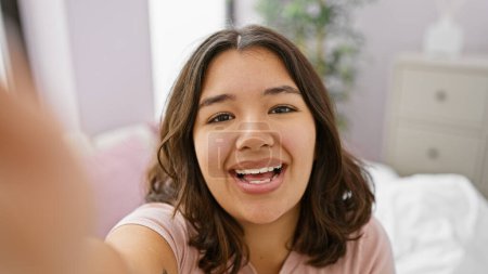 Téléchargez les photos : Une jeune femme hispanique joyeuse prend un selfie dans un cadre confortable de chambre à coucher, représentant la joie et la vie quotidienne. - en image libre de droit