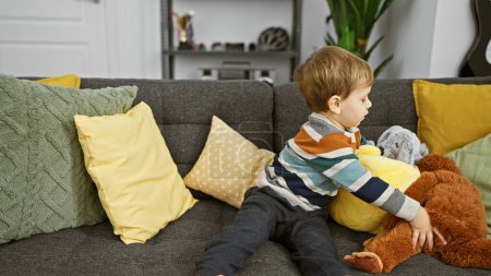 Téléchargez les photos : Jeune garçon blond relaxant sur un canapé avec des oreillers colorés et des jouets en peluche dans un salon confortable. - en image libre de droit