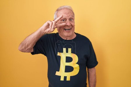 Téléchargez les photos : Homme âgé avec les cheveux gris portant Bitcoin t-shirt faisant symbole de paix avec les doigts sur le visage, souriant joyeux montrant la victoire - en image libre de droit