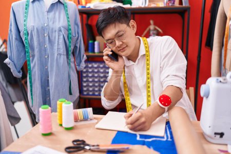 Téléchargez les photos : Jeune chinois tailleur parler sur smartphone écriture sur ordinateur portable à l'atelier - en image libre de droit