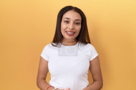 Photo for Young arab woman wearing casual white t shirt over yellow background with hands together and crossed fingers smiling relaxed and cheerful. success and optimistic - Royalty Free Image