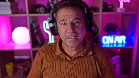 Foto de Hombre maduro con auriculares en una colorida sala de juegos iluminada sonríe a la cámara, proyectando ocio y tecnología en casa. - Imagen libre de derechos
