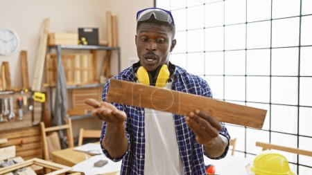 Photo for African american man examining wood in a carpentry workshop, wearing safety gear. - Royalty Free Image