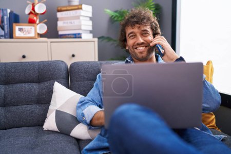 Téléchargez les photos : Young hispanic man talking on smartphone using laptop at home - en image libre de droit