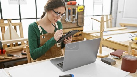 Photo for A woman wearing safety glasses and an apron taking notes on a digital tablet in a carpentry workshop with woodworking tools and a laptop on the desk. - Royalty Free Image