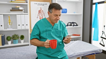 Mature man in scrubs sipping coffee and checking smartphone in a physiotherapy clinic interior.