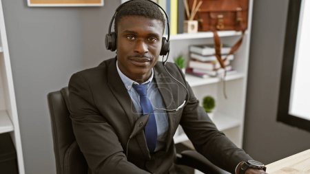 Photo for Handsome african man wearing a headset and suit seated in a modern office. - Royalty Free Image