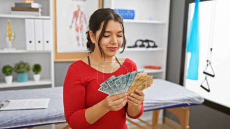 Photo for A smiling young woman examines brazilian reais in a bright rehabilitation clinic room. - Royalty Free Image