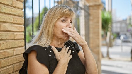 Caucasian blonde, middle age woman looking serious yet relaxed, standing outdoors in sunny city street coughing, showing signs of flu, possibly covid-19, indicating urban infection concern