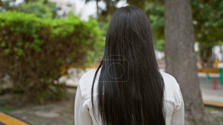Vue arrière d'une jeune femme hispanique dans un parc avec des arbres mettant en valeur la sérénité et la solitude en plein air urbaine.