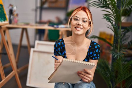 Foto de Joven mujer caucásica artista sonriendo dibujo seguro en el cuaderno en el estudio de arte - Imagen libre de derechos