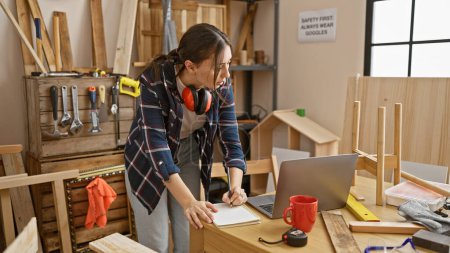 Téléchargez les photos : Une jeune femme focalisée dessine des dessins dans son atelier de menuiserie entouré d'outils et de bois. - en image libre de droit
