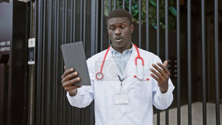 Photo for African male doctor with stethoscope and tablet outside a clinic communicating on a video call - Royalty Free Image