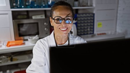 Foto de Una mujer hispana sonriente trabajando en una computadora en un laboratorio, exudando confianza y experiencia. - Imagen libre de derechos