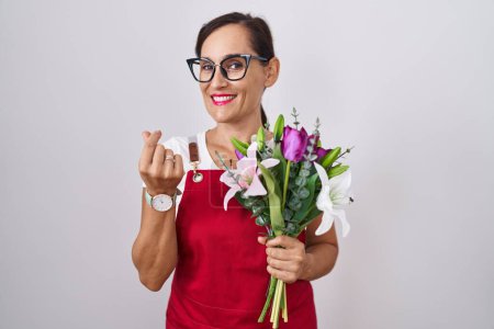 Téléchargez les photos : Femme brune d'âge moyen portant un tablier travaillant au magasin de fleuristes tenant un bouquet faisant un geste d'argent avec les mains, demandant un paiement de salaire, entreprise millionnaire - en image libre de droit