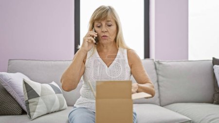 Upset middle-aged blonde woman unpacking a cardboard box at home, engrossed in a serious phone conversation